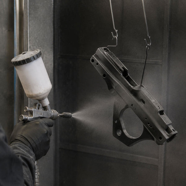 Person spraying Cerakote on a black metal object with a spray gun in a controlled environment.