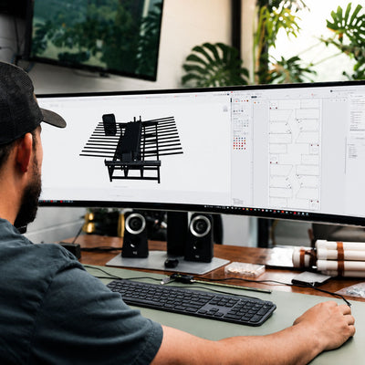 Engineer working at a desk with dual monitors, one displaying a 3D model of a machine.